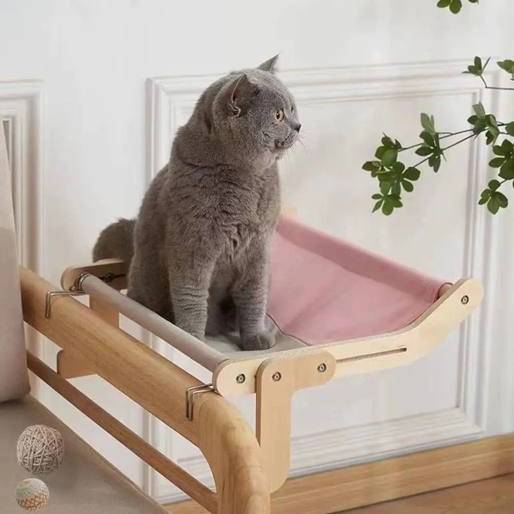 Gray cat sitting on a pink canvas-lined window-mounted hammock bed with a wooden frame, designed for comfort and elevated views.